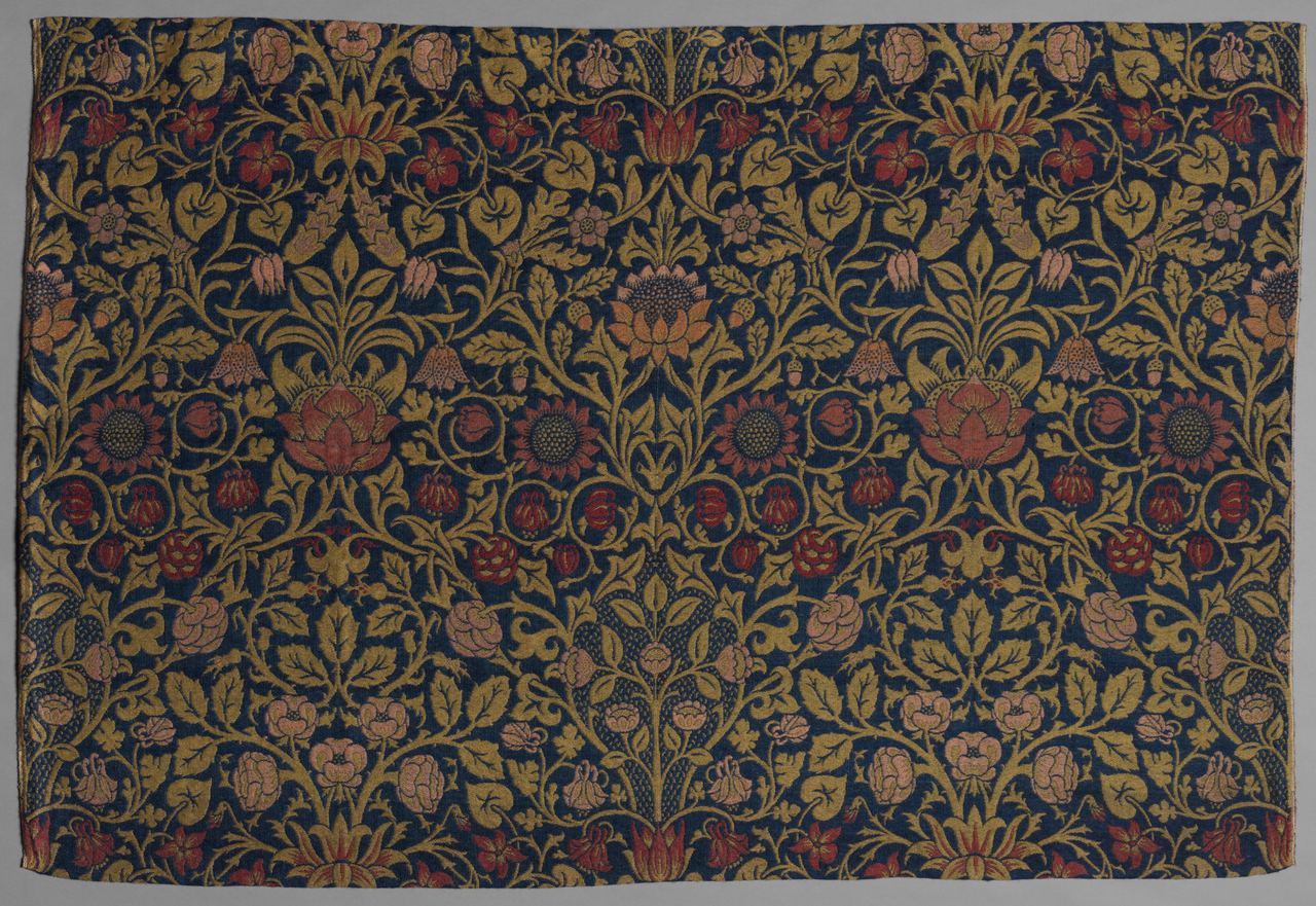 A dark blue woven textile covered in a dense, symmetrical pattern of gold-green vines and leaves with red, pink and orange flowers including sunflowers, columbines and violets. The pattern repeats across the width of the cloth.