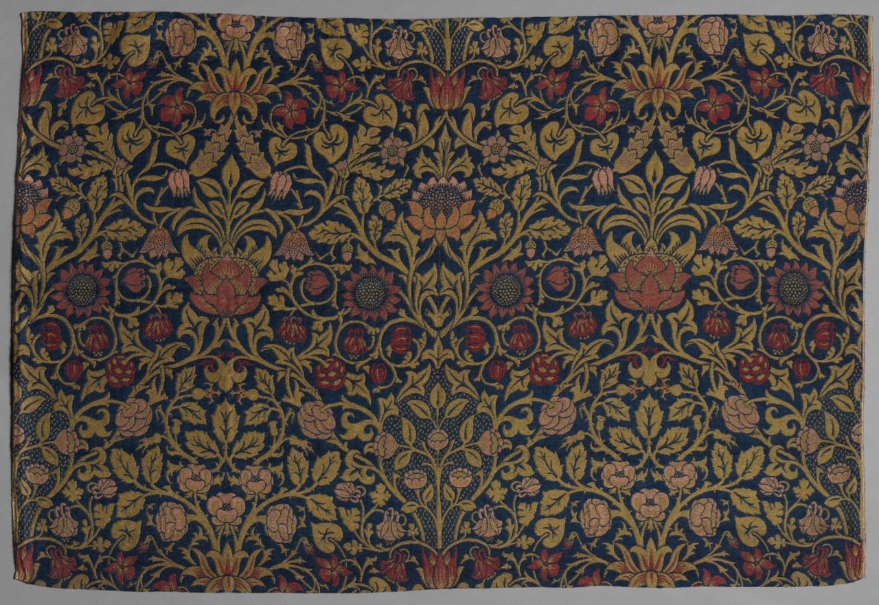 A dark blue woven textile covered in a dense, symmetrical pattern of gold-green vines and leaves with red, pink and orange flowers including sunflowers, columbines and violets. The pattern repeats across the width of the cloth.