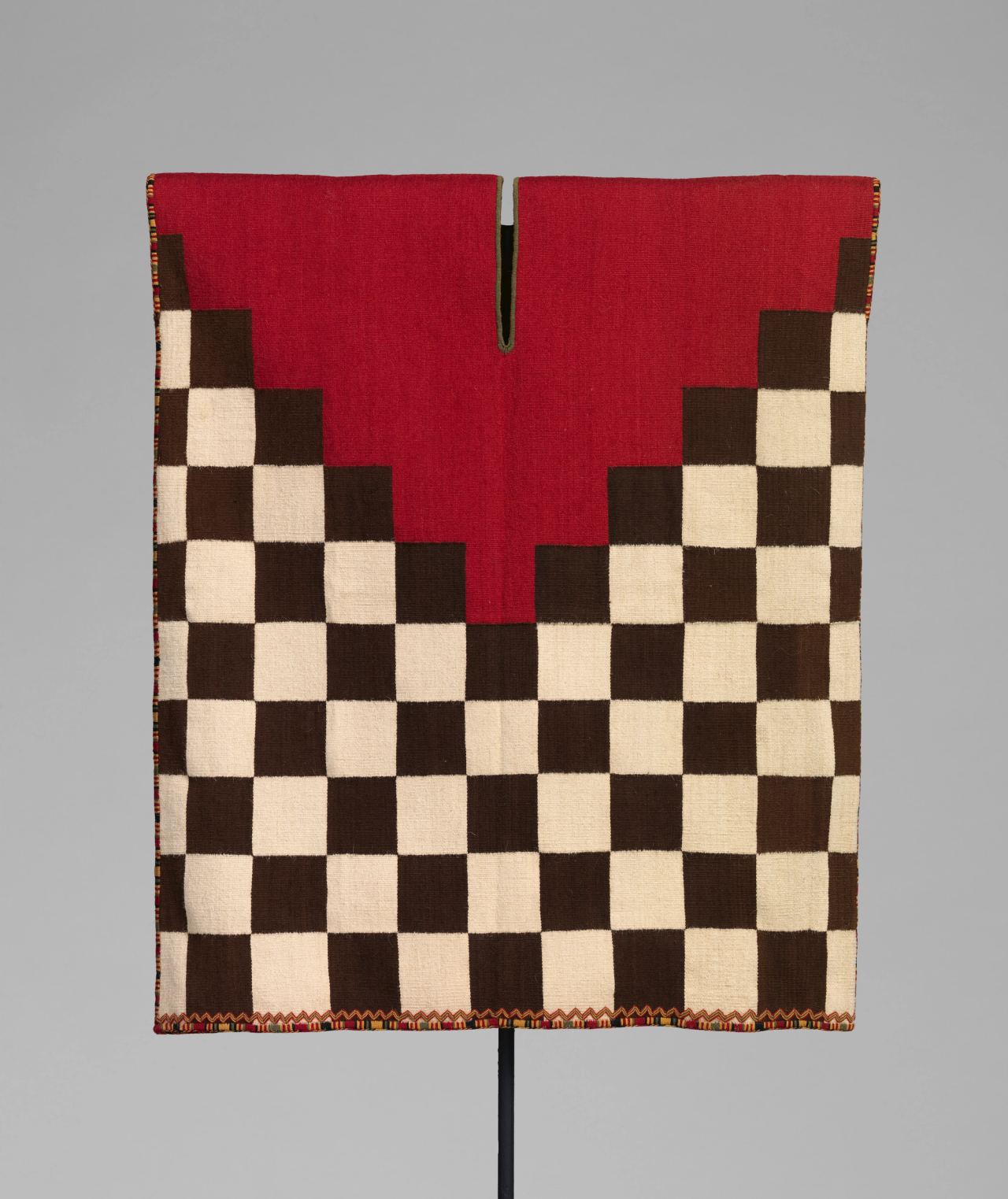 A miniature checkerboard tunic in cream and brown squares with a red V-shaped yoke, shown flat.