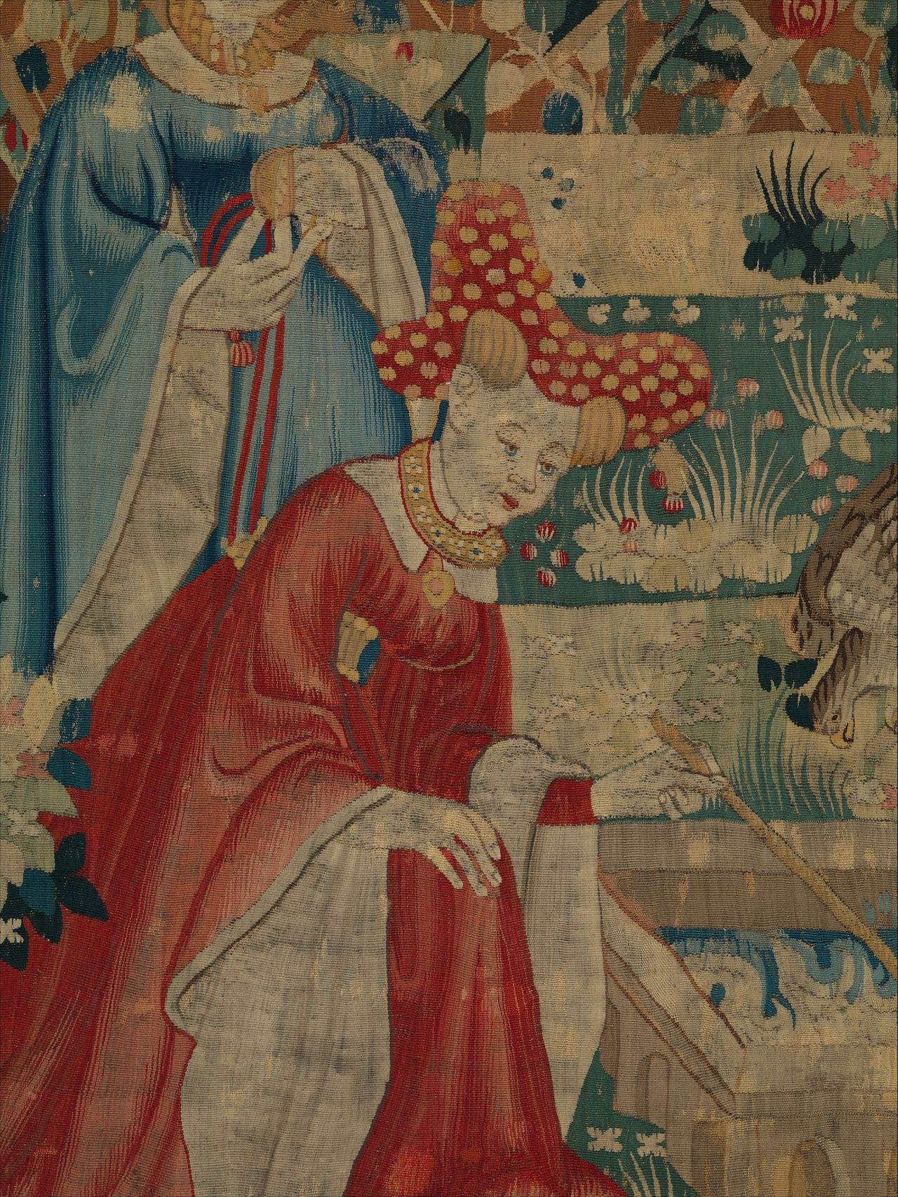 Close view of woven wool: a woman in a red gown and a tall red hat dotted with yellow bends over at the center, with a figure in blue behind her. The surface is finely ribbed, and where one color meets another the edges step and interlock in small teeth instead of following a line of thread laid on top.