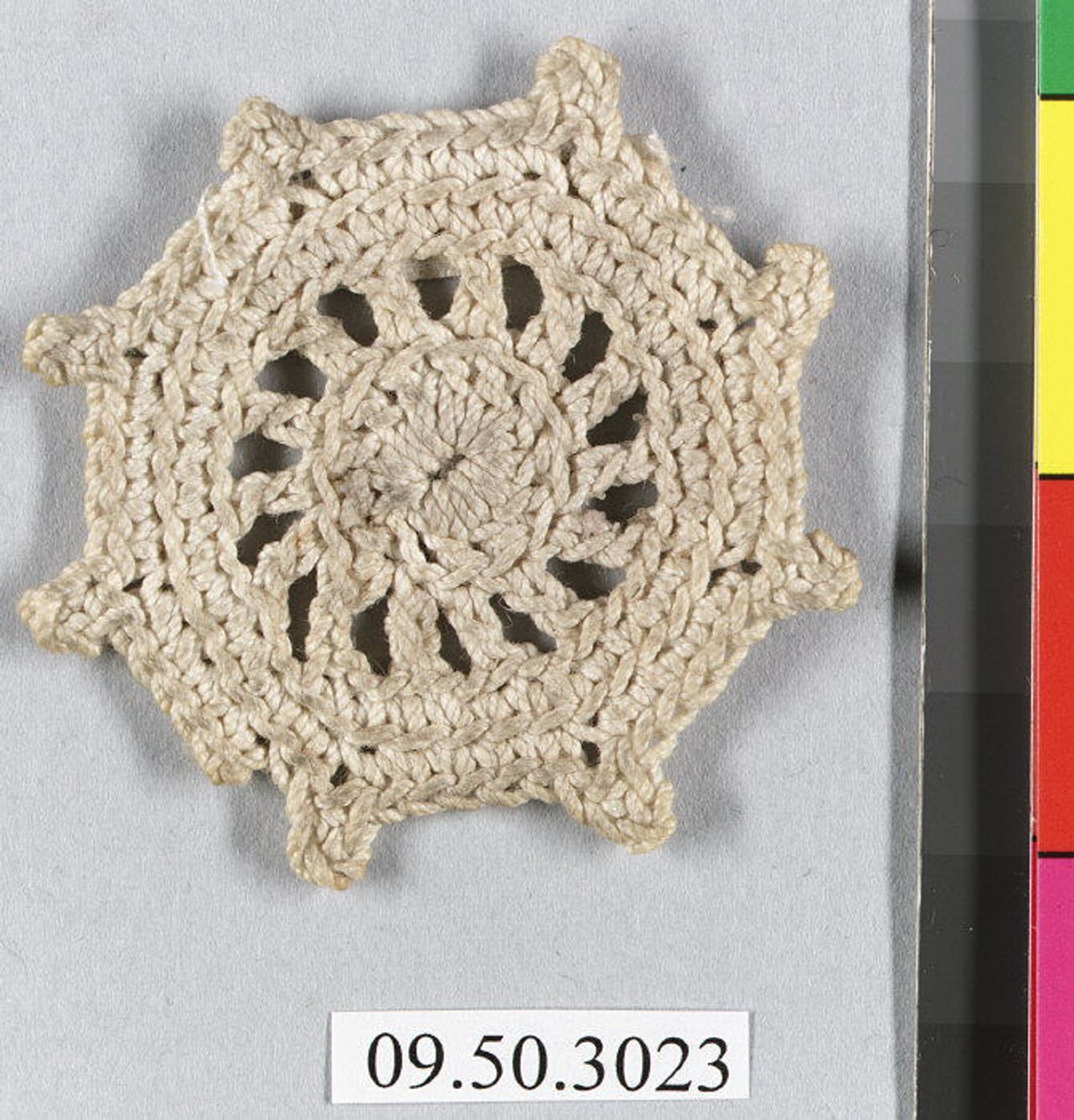 A small round cream-colored crochet motif worked outward from a solid center, with a ring of open spokes inside and eight small projecting lobes around the edge.