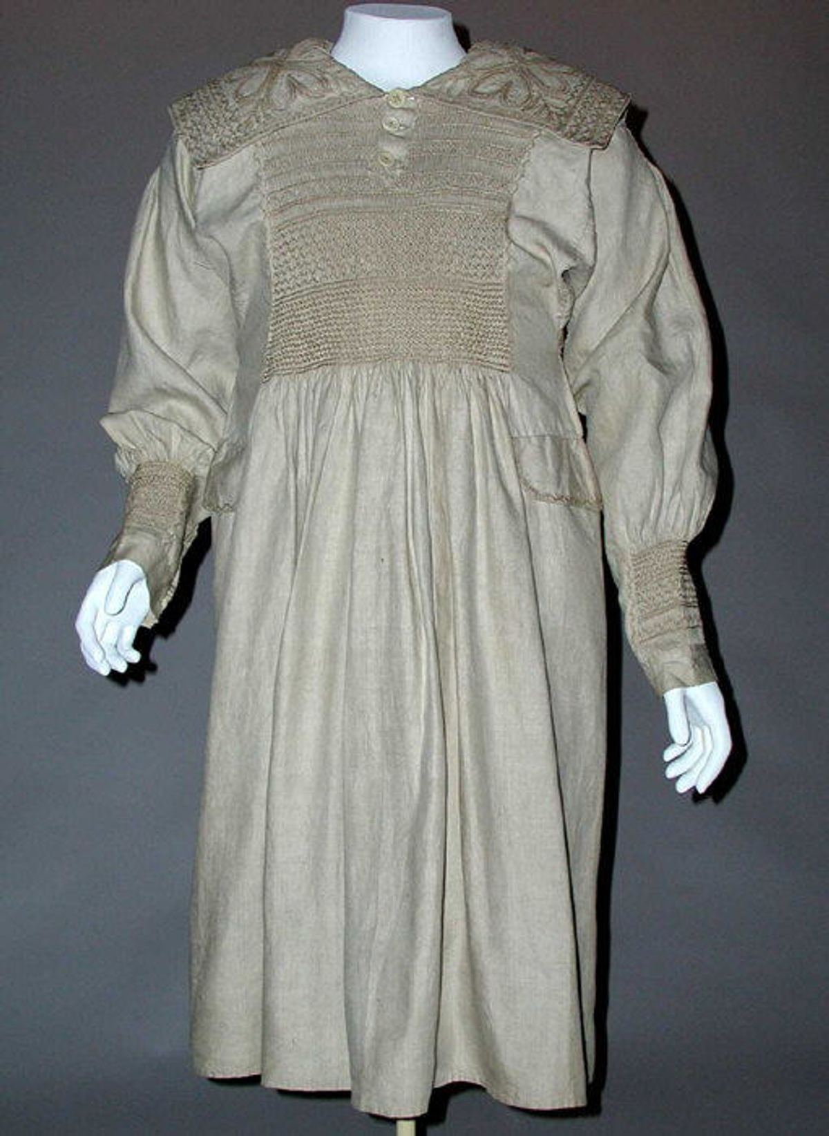 A man's linen smock of the mid to late 19th century on a mannequin, with a wide embroidered collar and a panel of smocking across the chest above a full gathered skirt.
