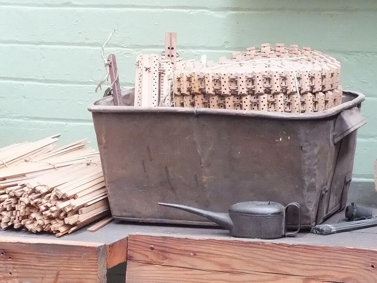 A rusty metal tub holding a stack of small wooden lags, each a short block pierced with rows of holes and some with pegs, linked together in a chain. Loose wooden strips lie in a bundle to the left and a black oil can sits in front, against a pale green brick wall.