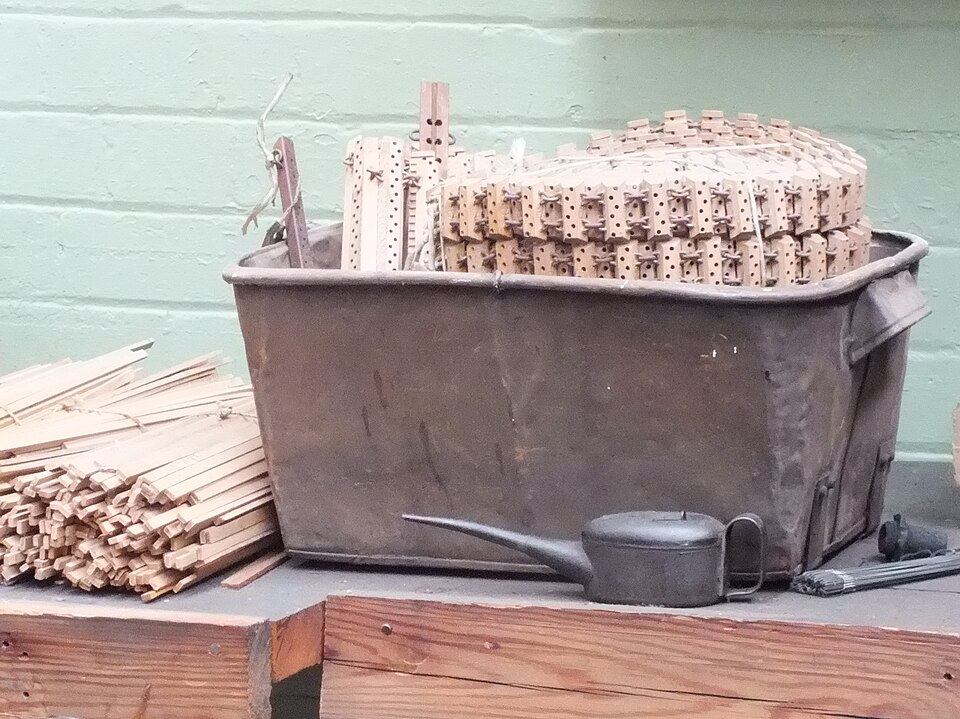 A rusty metal tub holding a stack of small wooden lags, each a short block pierced with rows of holes and some with pegs, linked together in a chain. Loose wooden strips lie in a bundle to the left and a black oil can sits in front, against a pale green brick wall.