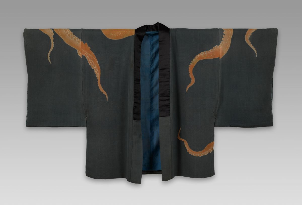 A short Japanese jacket laid flat with wide sleeves, in dark gray-green with a black satin collar band and a blue lining, orange octopus tentacles with rows of round suckers printed across the sleeves, shoulders and lower front.