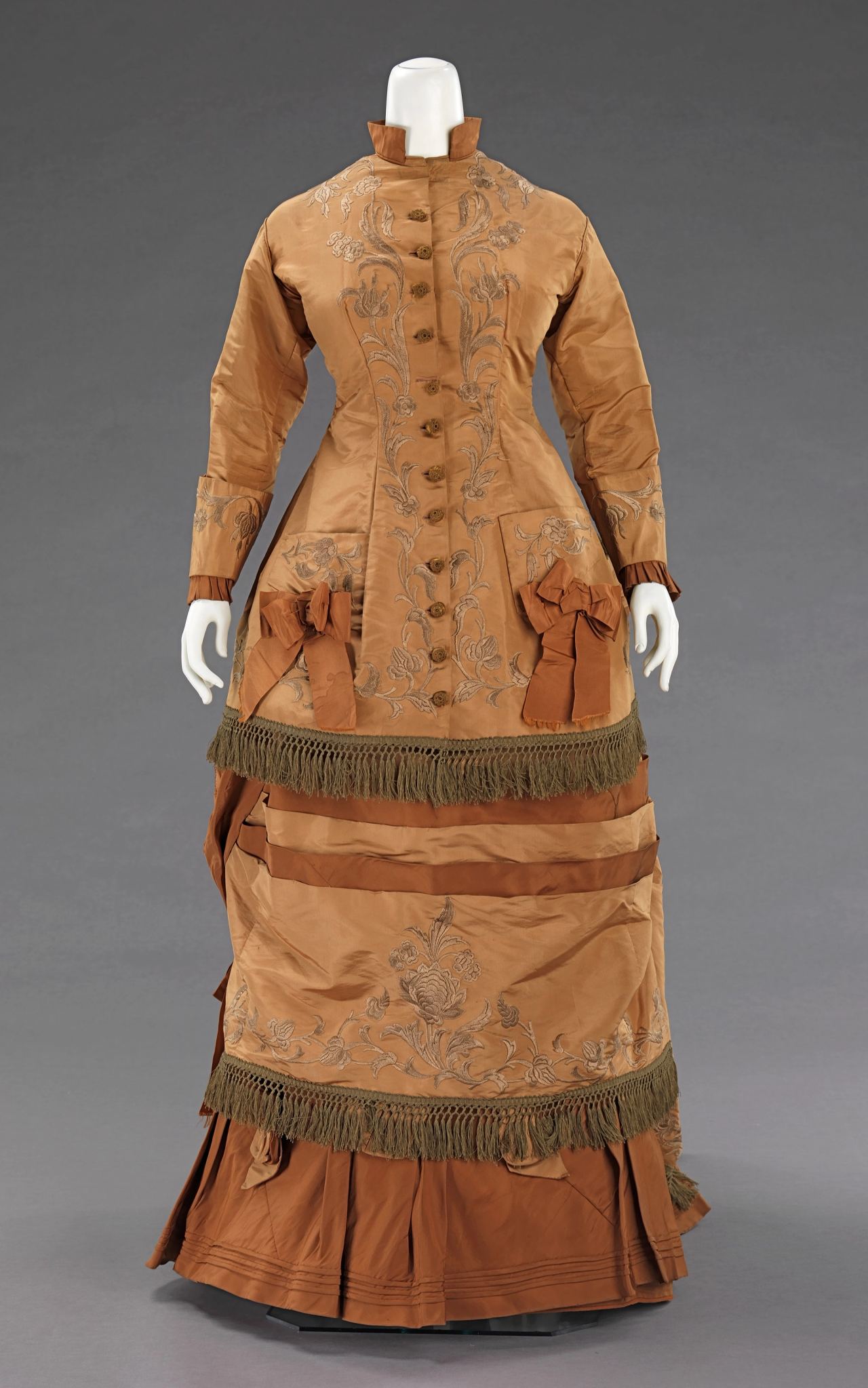 A woman's tan silk afternoon dress on a mannequin, its high-collared bodice closing with a row of small buttons down the center front, with embroidered floral scrolls, fringe, and ribbon bows on the overskirt.