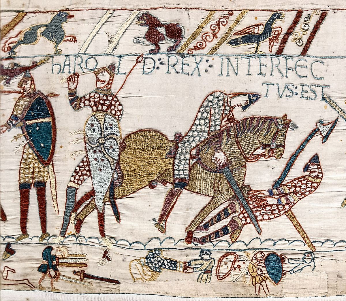 Detail of an embroidered cloth. A knight in a pointed helmet and mail falls forward over a horse whose body is filled with close rows of stitches; beside him another warrior raises a spear. A Latin inscription in blue-gray stitched letters runs above them, and the bare pale linen shows between the figures and along the border lines.