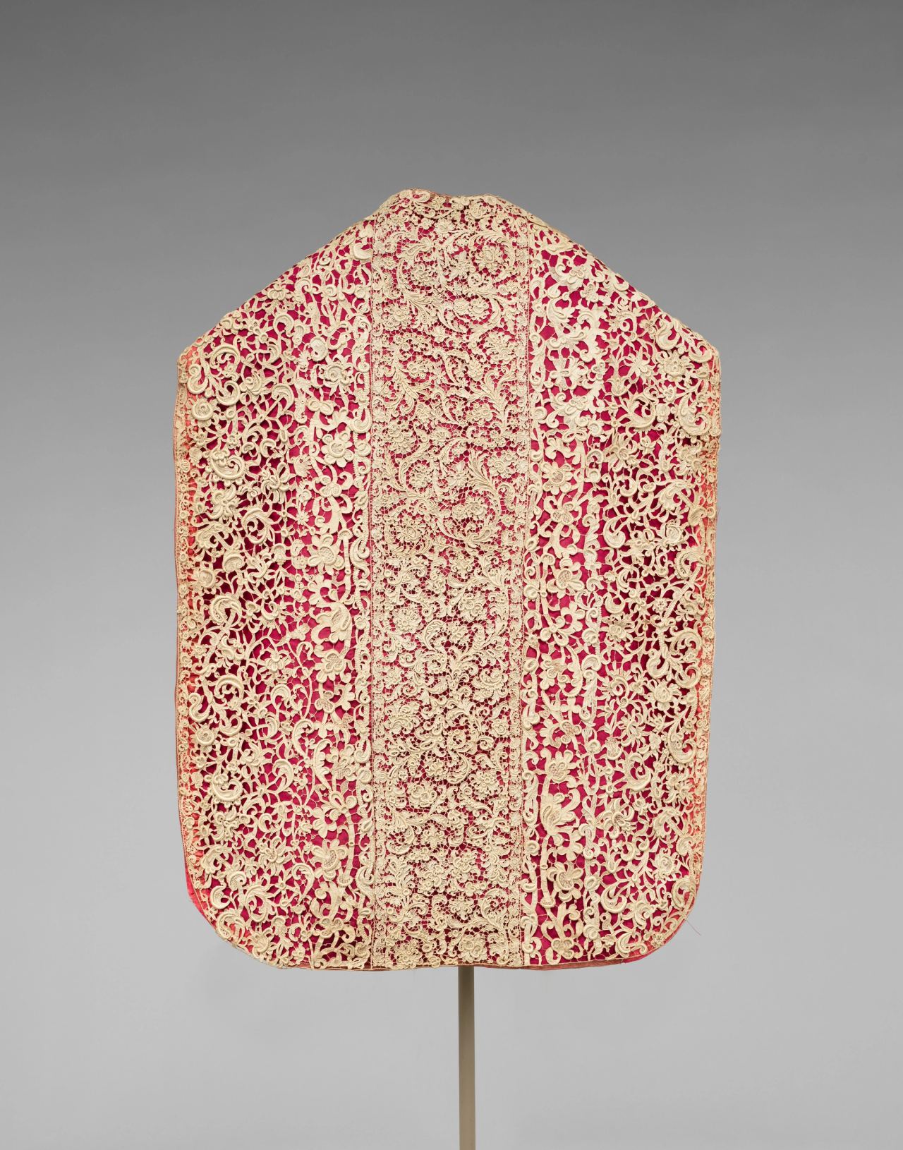 A sleeveless priest's chasuble seen flat against a gray backdrop: cream needle lace of scrolling flowers laid over bright red fabric, with a wide central vertical band of finer pattern between two broader side panels.