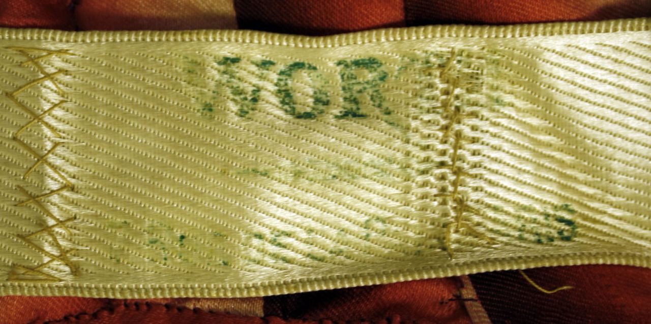 A cream ribbon label stamped in green ink, the lettering partly worn so that only part of the name can be read, held to the dress by hand cross stitches at the left and a short row of stitches at the right.