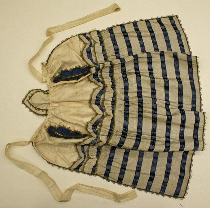 A cream silk apron laid flat, with blue and silver-coloured stripes, a bib-like top section with two lace-edged blue panels, and long cream ties.