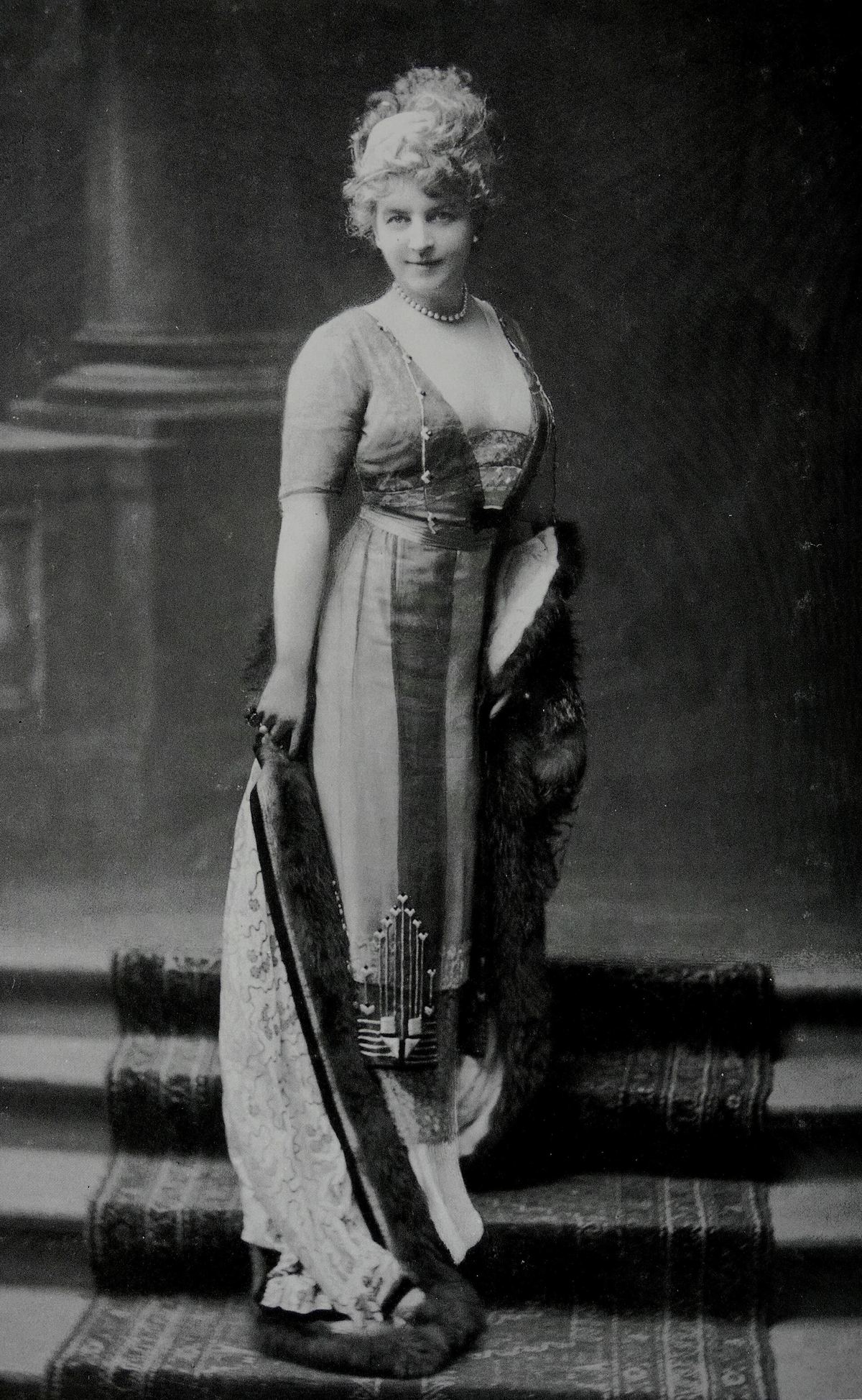 Black-and-white studio portrait of Jeanne Paquin standing on a carpeted stair in a long, close-fitting evening dress with a sheer overlay, a deep V neckline and a dark geometric embroidered motif at the hem, a fur stole held across her arm and a string of pearls at her neck.