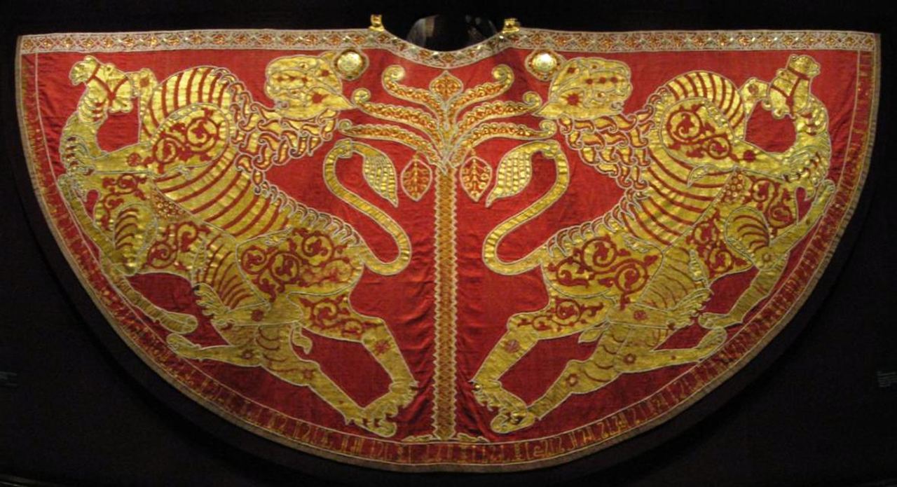 A semicircular red mantle laid flat against a dark ground. Two gold lions, embroidered in profile, face each other across a slender central tree, and a narrow border of small motifs runs along the curved lower edge.