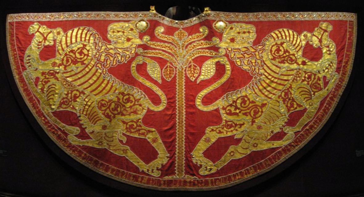 A semicircular red mantle laid flat against a dark ground. Two gold lions, embroidered in profile, face each other across a slender central tree, and a narrow border of small motifs runs along the curved lower edge.