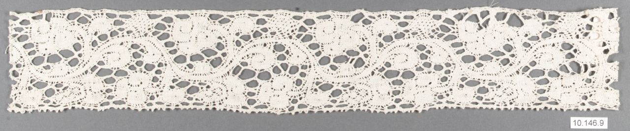 A narrow strip of cream bobbin lace. Scrolling stems and solid leaf-like patches are separated by irregular open holes, with a small toothed edge along the bottom, shown against a gray background.