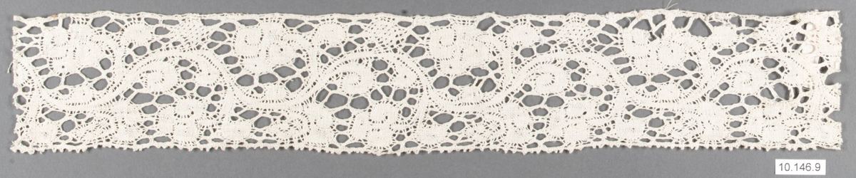 A narrow strip of cream bobbin lace. Scrolling stems and solid leaf-like patches are separated by irregular open holes, with a small toothed edge along the bottom, shown against a gray background.