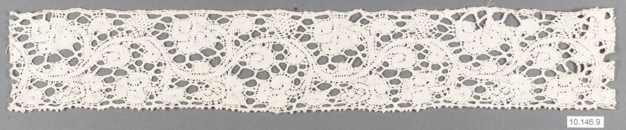 A narrow strip of cream bobbin lace. Scrolling stems and solid leaf-like patches are separated by irregular open holes, with a small toothed edge along the bottom, shown against a gray background.