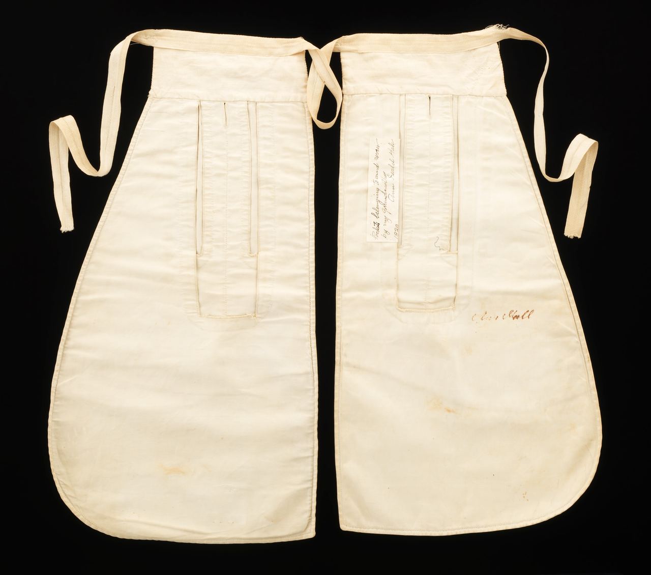 A pair of white tie-on pockets laid flat on black, each with a long vertical opening near the top and a waist tape; handwriting in ink is visible on a paper label and on the lower part of the right-hand pocket.