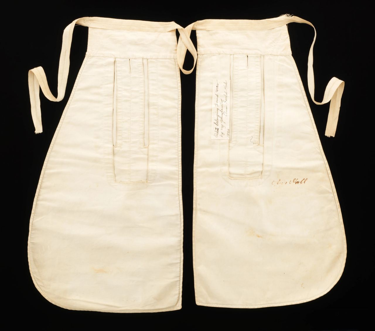 A pair of white tie-on pockets laid flat on black, each with a long vertical opening near the top and a waist tape; handwriting in ink is visible on a paper label and on the lower part of the right-hand pocket.