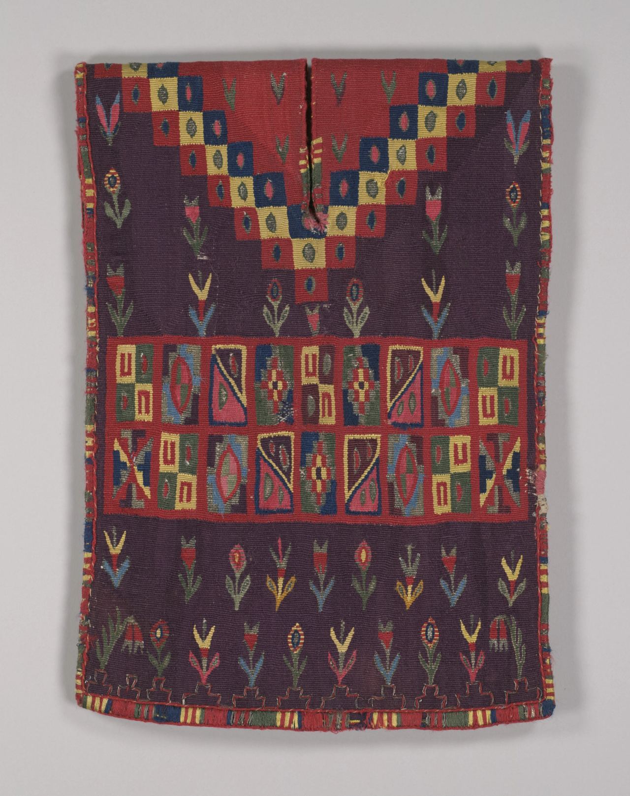 A small purple garment with a red yoke of stepped yellow and blue checks, a central band of boxed geometric motifs and rows of small flowers, shown flat.