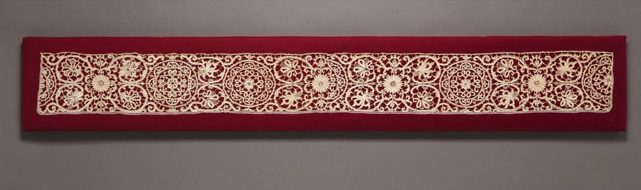 A long, narrow strip of cream needle lace mounted on red cloth: round and flower-like medallions alternate with sprays of leaves along the whole length, with a straight, finely toothed edge.