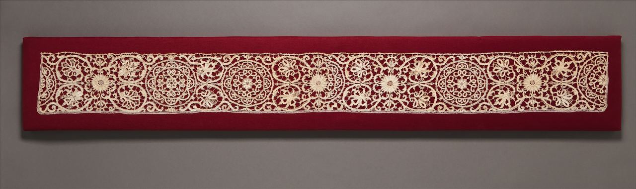 A long, narrow strip of cream needle lace mounted on red cloth: round and flower-like medallions alternate with sprays of leaves along the whole length, with a straight, finely toothed edge.