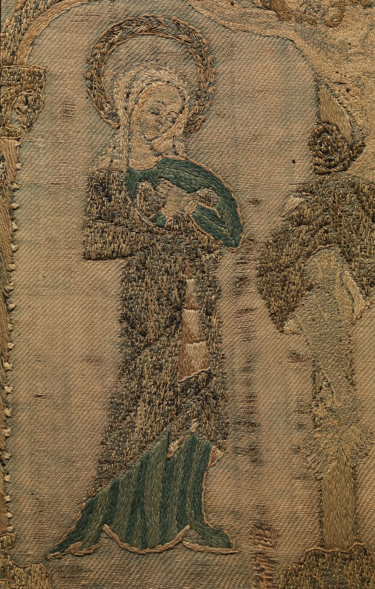 Close view of a small standing saint with a halo, stitched in green, cream and gold-colored thread. A plain woven ground with a fine diagonal rib shows all around the figure, and individual stitches can be seen lying over it.