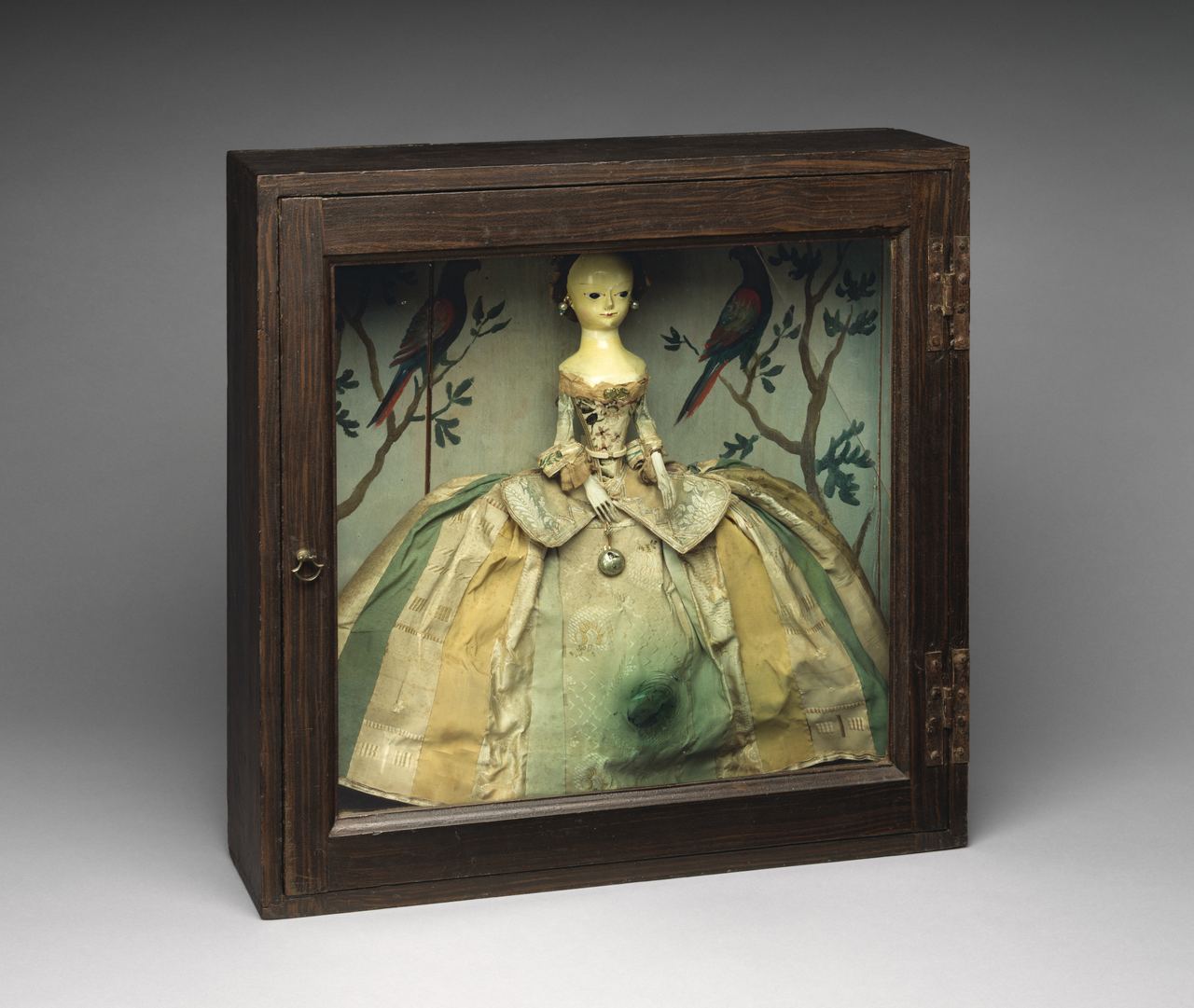 The same doll seen through the glass front of her closed box: pearl earrings, a pocket watch hanging at her waist, and a skirt of striped silks.