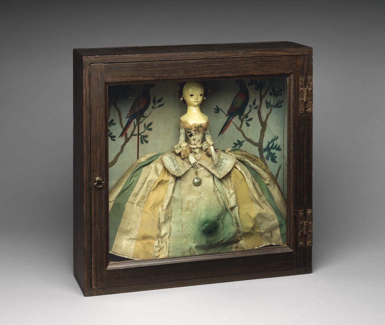 The same doll seen through the glass front of her closed box: pearl earrings, a pocket watch hanging at her waist, and a skirt of striped silks.