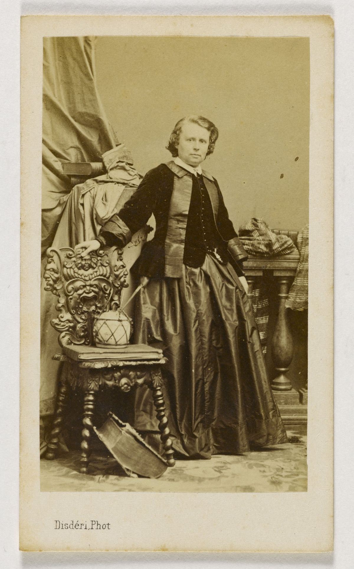 Carte-de-visite photograph of Rosa Bonheur standing full length beside a carved wooden chair, one hand resting on its back. She wears a dark velvet jacket with lighter silk panels and a white collar over a long, full silk skirt, and her wavy hair falls to jaw length. The name Disdéri is printed on the card mount.