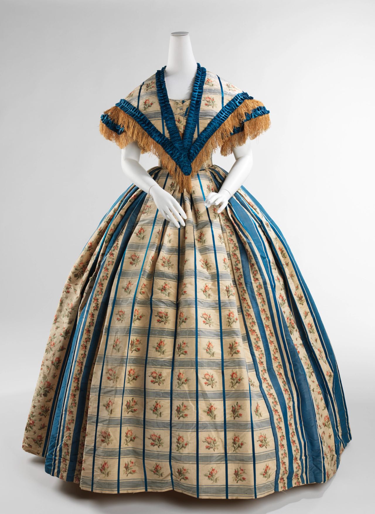 A full-skirted evening dress on a mannequin: cream silk woven with small floral sprigs in a plaid of blue stripes, an off-the-shoulder V-shaped cape collar edged with blue ruching and gold fringe, and a very wide bell-shaped skirt.