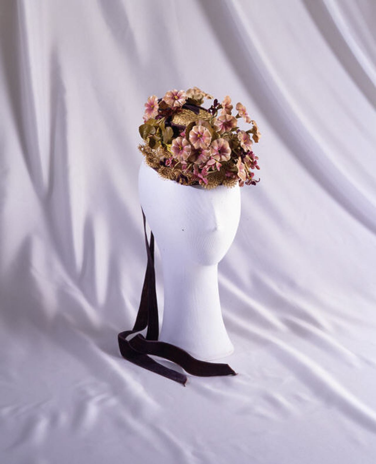 A small hat on a white mannequin head, covered with a crown of pink and cream artificial blossoms and green leaves, with long dark ribbons trailing down on either side.