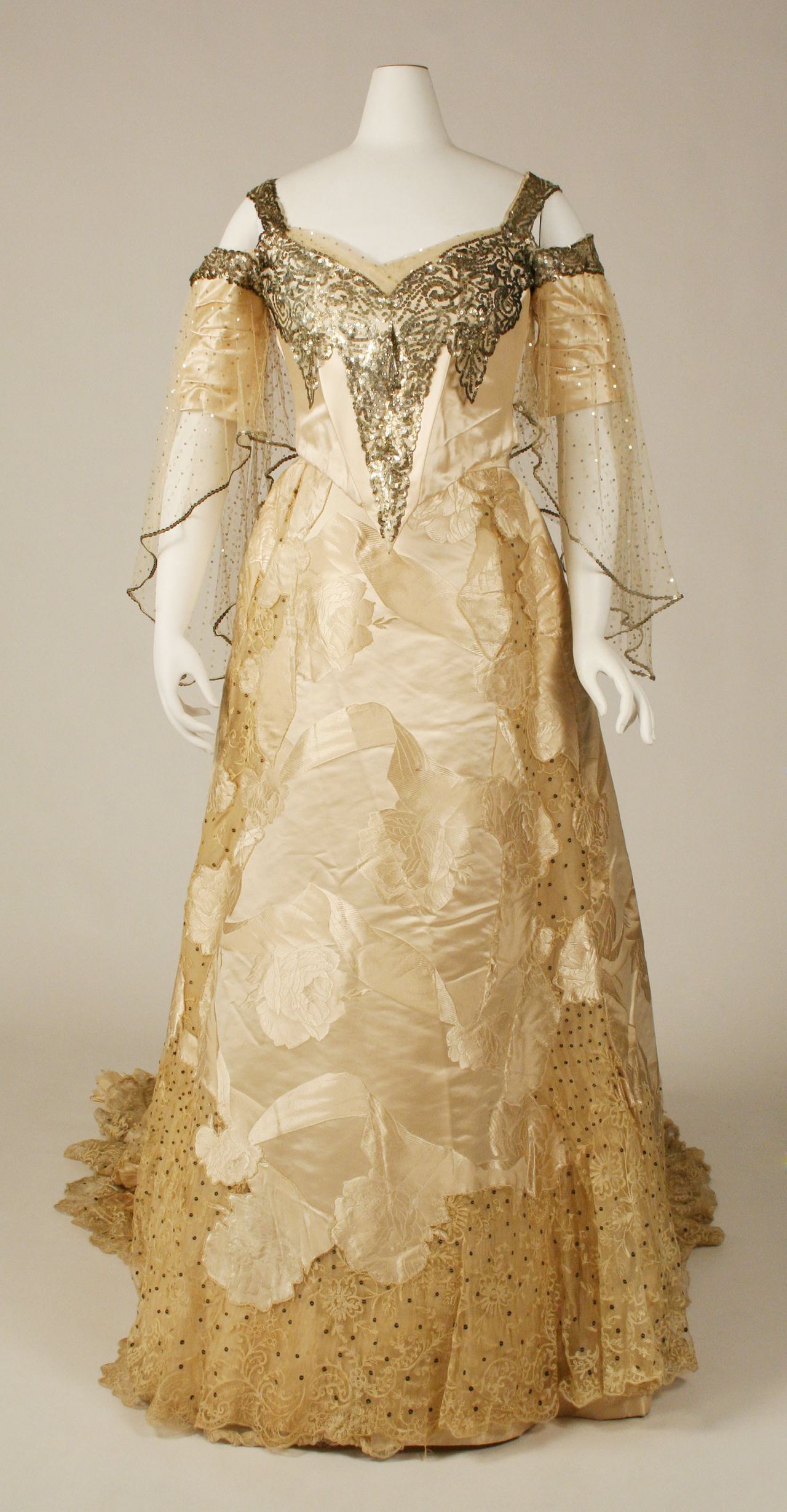 A cream satin ball gown with a long train on a mannequin, its pointed bodice and off-shoulder net sleeves glittering with sequins, and sequin-dotted net around the hem.