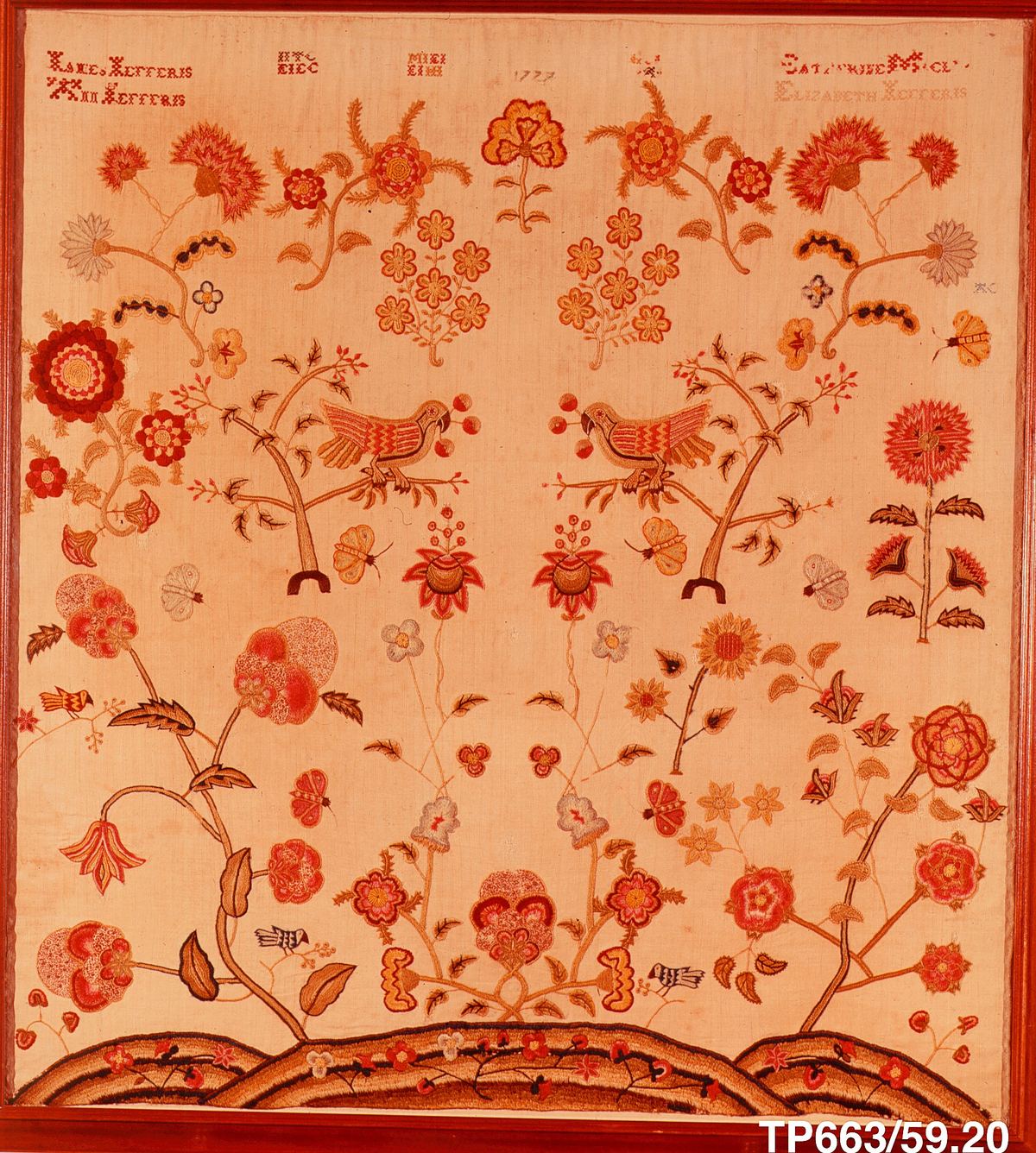 A symmetrical embroidered picture on pale linen: two parrots on small trees at the center, large flowers, butterflies and birds around them, and a band of rolling hills at the bottom. Rows of tiny red crossed stitches spell names and the date 1777 across the top.
