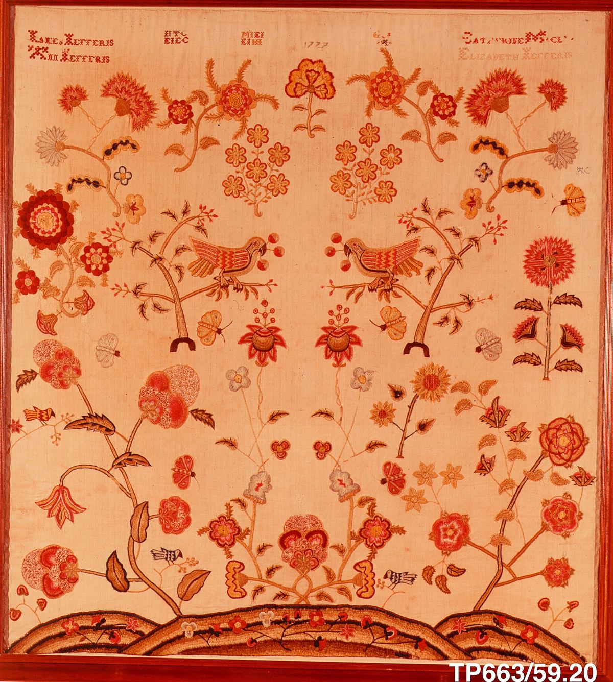 A symmetrical embroidered picture on pale linen: two parrots on small trees at the center, large flowers, butterflies and birds around them, and a band of rolling hills at the bottom. Rows of tiny red crossed stitches spell names and the date 1777 across the top.