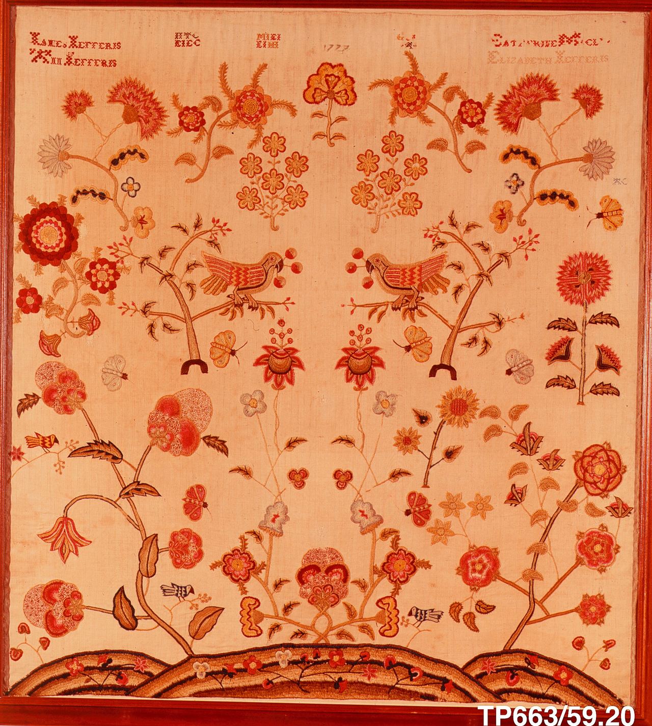 A symmetrical embroidered picture on pale linen: two parrots on small trees at the center, large flowers, butterflies and birds around them, and a band of rolling hills at the bottom. Rows of tiny red crossed stitches spell names and the date 1777 across the top.