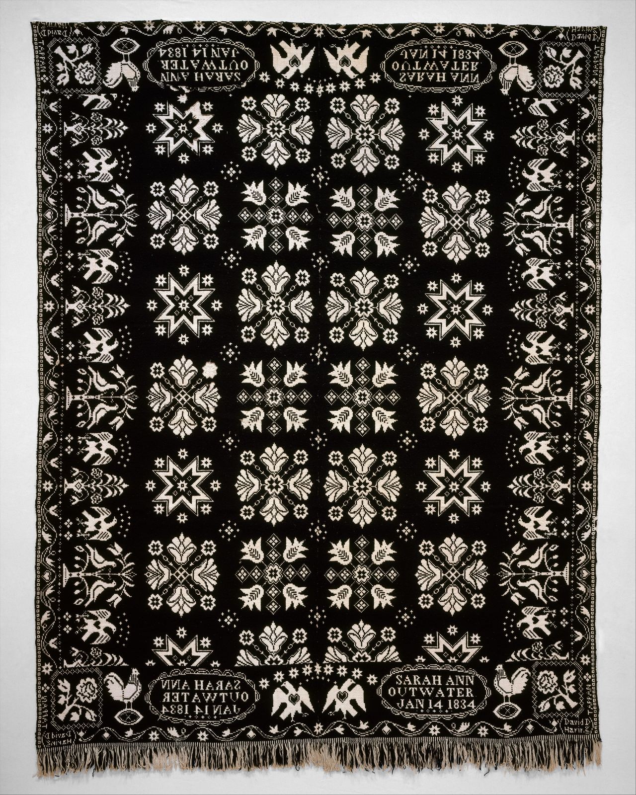 A black and white woven coverlet in a symmetrical layout. A grid of white star and flower motifs fills the center, framed by a border of birds and flowering plants. Oval panels in the four corners are lettered Sarah Ann Outwater, Jan 14 1834, and fringe hangs along the bottom edge.