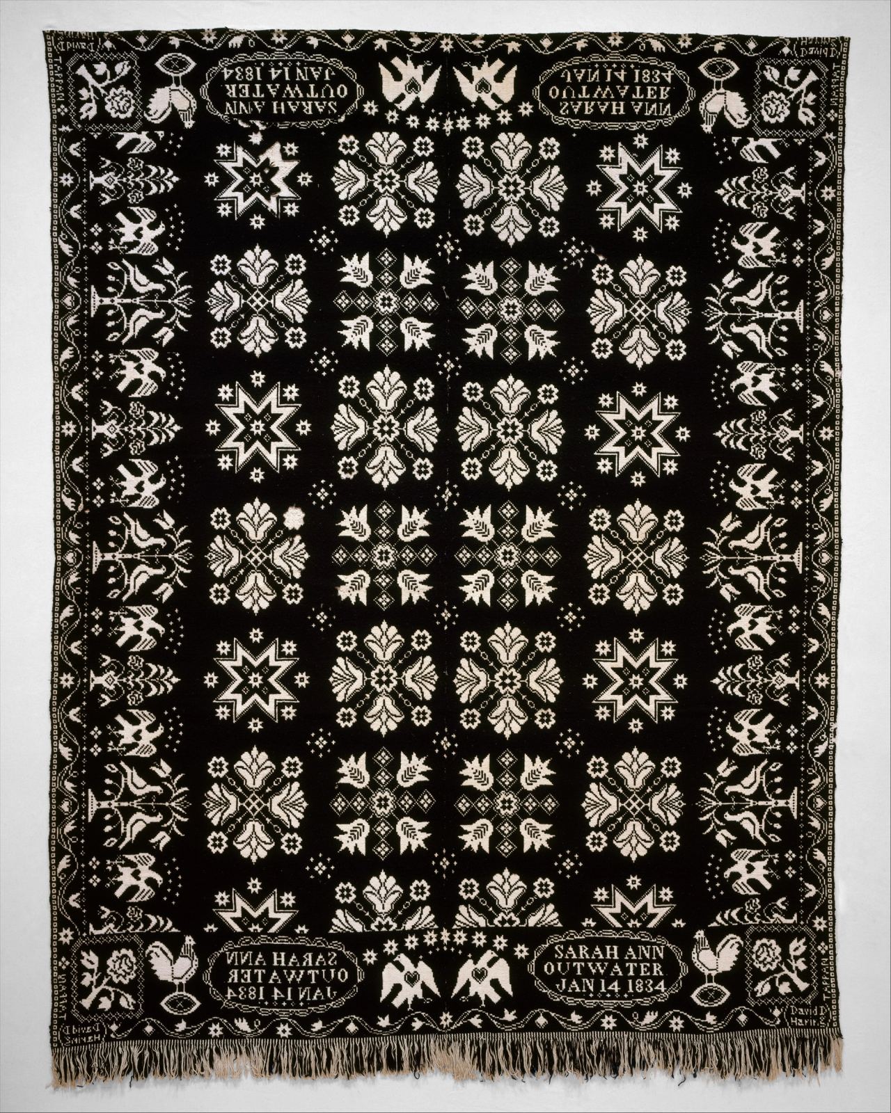 A black and white woven coverlet in a symmetrical layout. A grid of white star and flower motifs fills the center, framed by a border of birds and flowering plants. Oval panels in the four corners are lettered Sarah Ann Outwater, Jan 14 1834, and fringe hangs along the bottom edge.