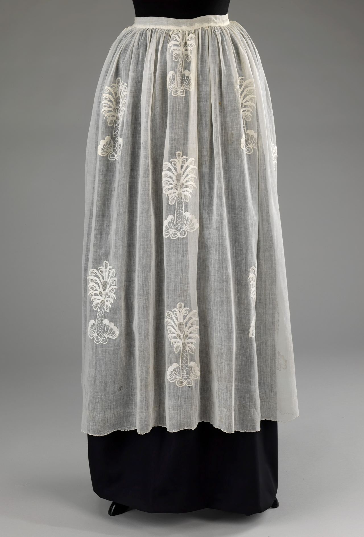 A sheer white apron gathered at the waist and worn over a dark skirt on a mannequin, with white embroidered tree-like motifs scattered across the gauzy cotton.