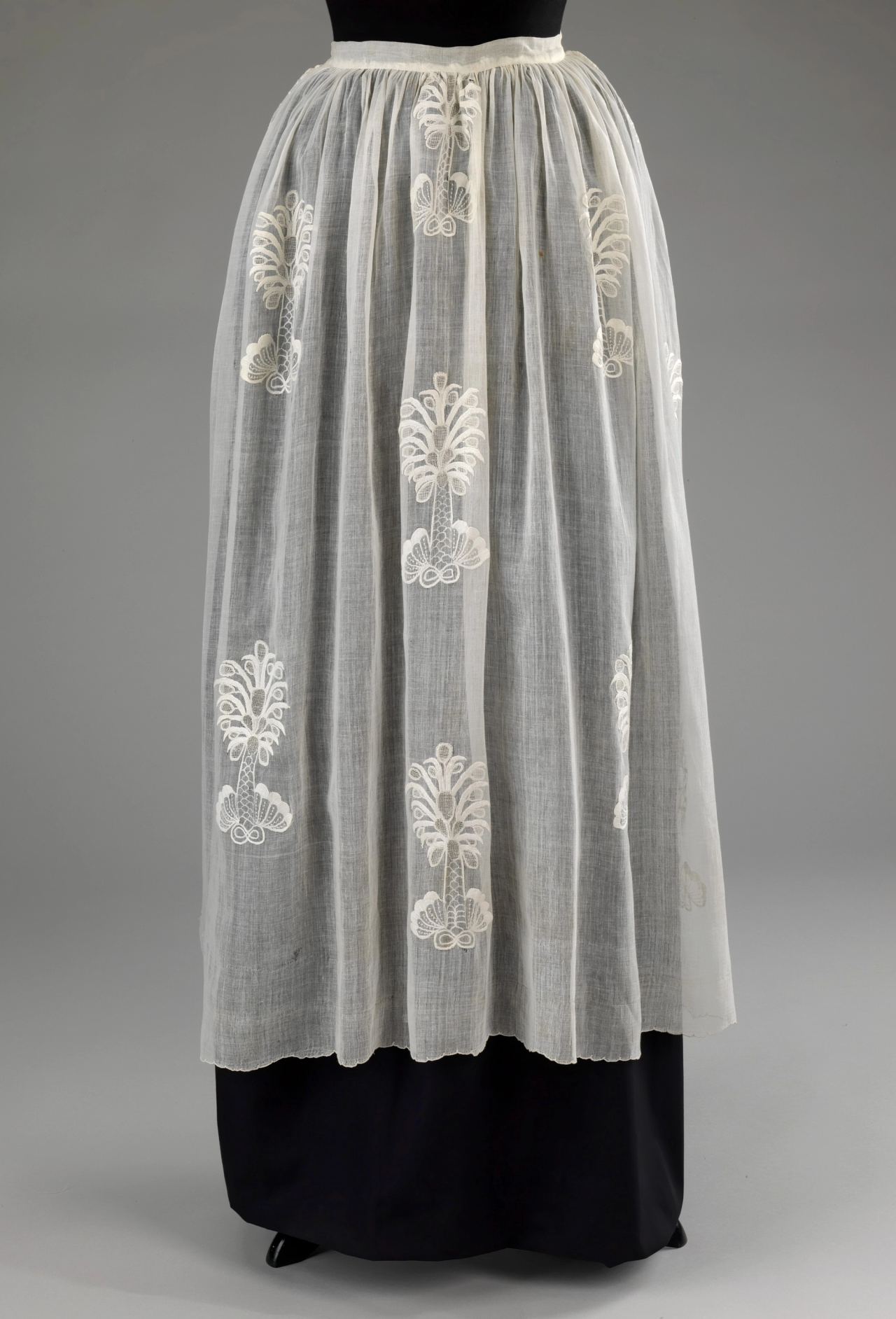 A sheer white apron gathered at the waist and worn over a dark skirt on a mannequin, with white embroidered tree-like motifs scattered across the gauzy cotton.