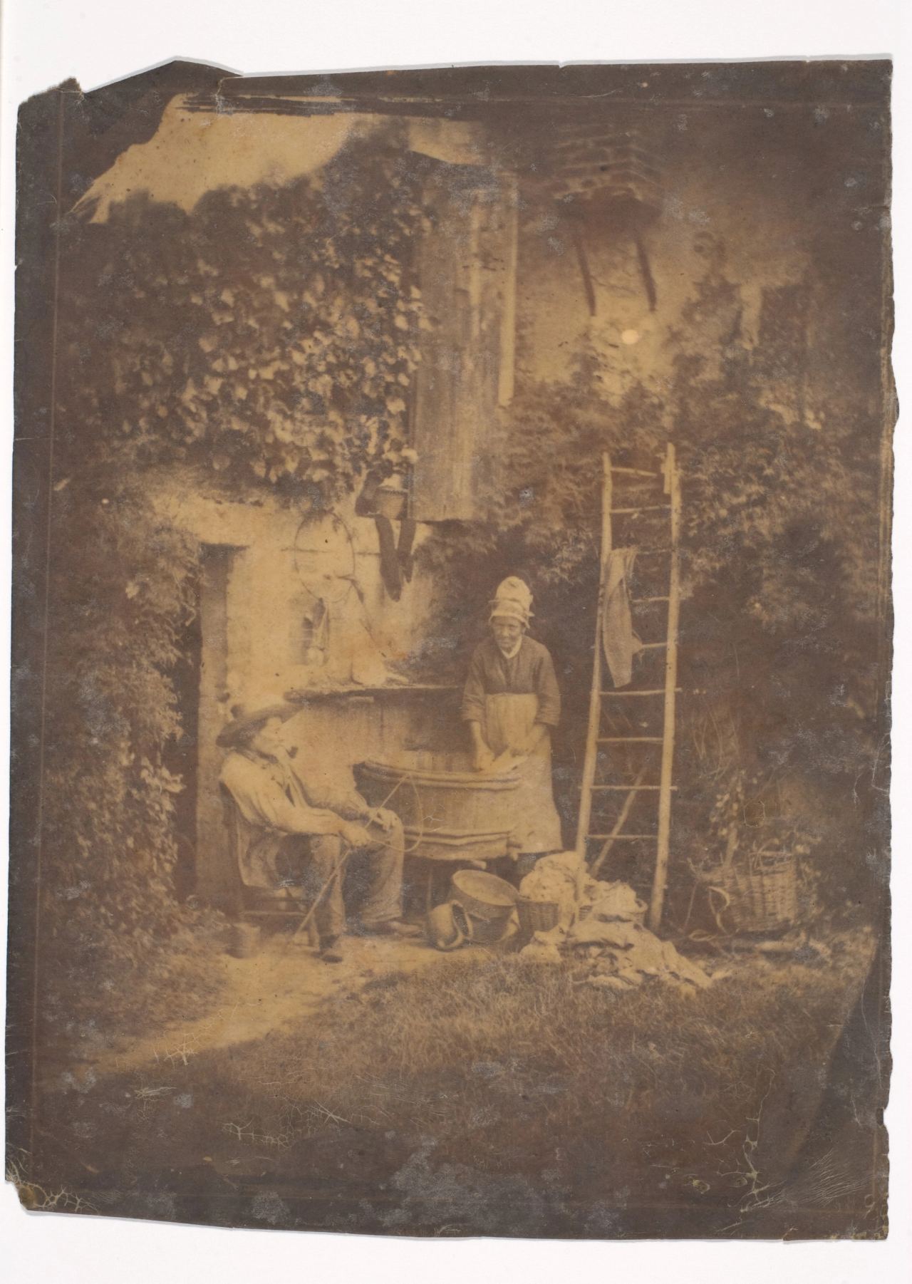 A woman in a white cap and apron stands at a large wooden tub, her hands in the washing, beside a seated man in a hat holding a stick; a ladder with a garment hung over it leans on the wall, with baskets and a heap of cloth on the grass.