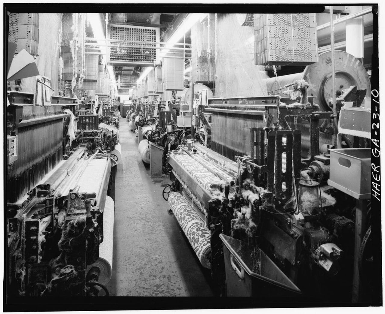 Black and white photograph down an aisle between rows of large looms in a textile mill. At upper right a tall stack of rectangular cards punched with small holes hangs above a loom, and bands of warp threads run down from the machinery overhead.