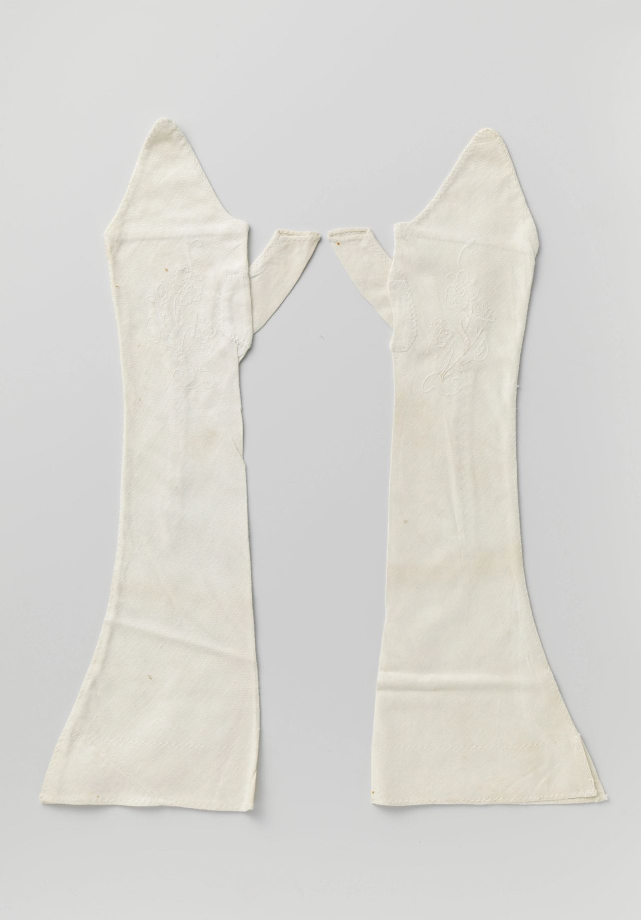 A pair of white cotton mitts laid flat on a grey ground, each long and widening toward the cuff with a separate thumb piece and faint white floral embroidery on the back of the hand.