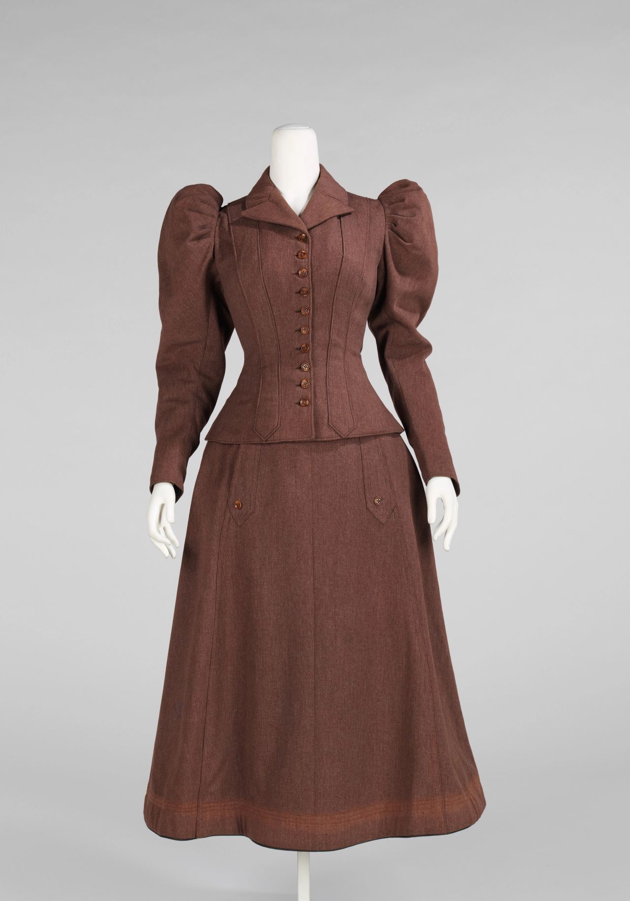 A reddish-brown wool suit on a white mannequin: a fitted jacket with very large puffed sleeves, a row of buttons and a pointed collar, and a plain flared skirt that ends well above the floor, with rows of stitching at the hem.