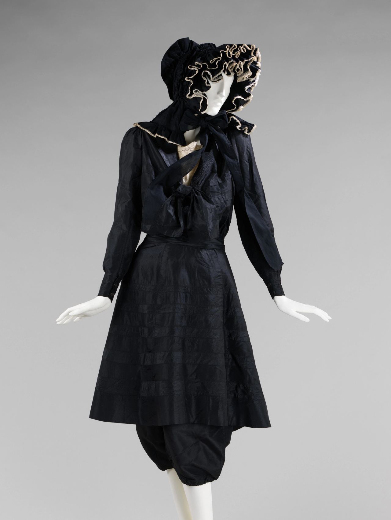 A black silk bathing costume on a mannequin: a blouse with a large bow, a knee-length skirt with horizontal bands of stitching, a ruffled bonnet edged in cream lace, and full gathered trousers visible beneath the skirt.