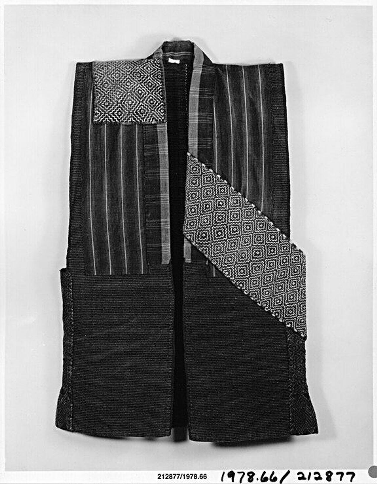 Black-and-white photograph of a sleeveless jacket pieced from many dark cloths: striped panels, a plain lower section, a square patch of diamond pattern at the shoulder and a wide diagonal band of diamond pattern across the front.