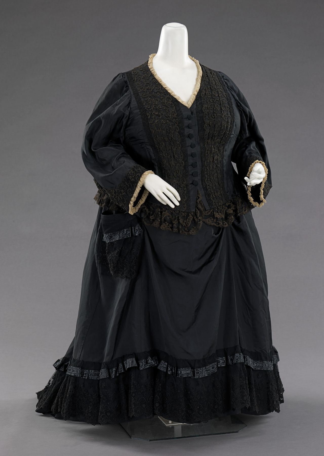 A woman's British mourning dress of 1894 on a mannequin: a black silk bodice and skirt with black lace panels, ribbon bands and ruffles, and a narrow cream ruche at the neck and cuffs.