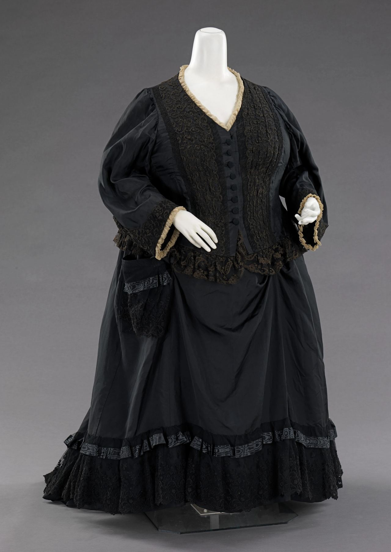A woman's British mourning dress of 1894 on a mannequin: a black silk bodice and skirt with black lace panels, ribbon bands and ruffles, and a narrow cream ruche at the neck and cuffs.