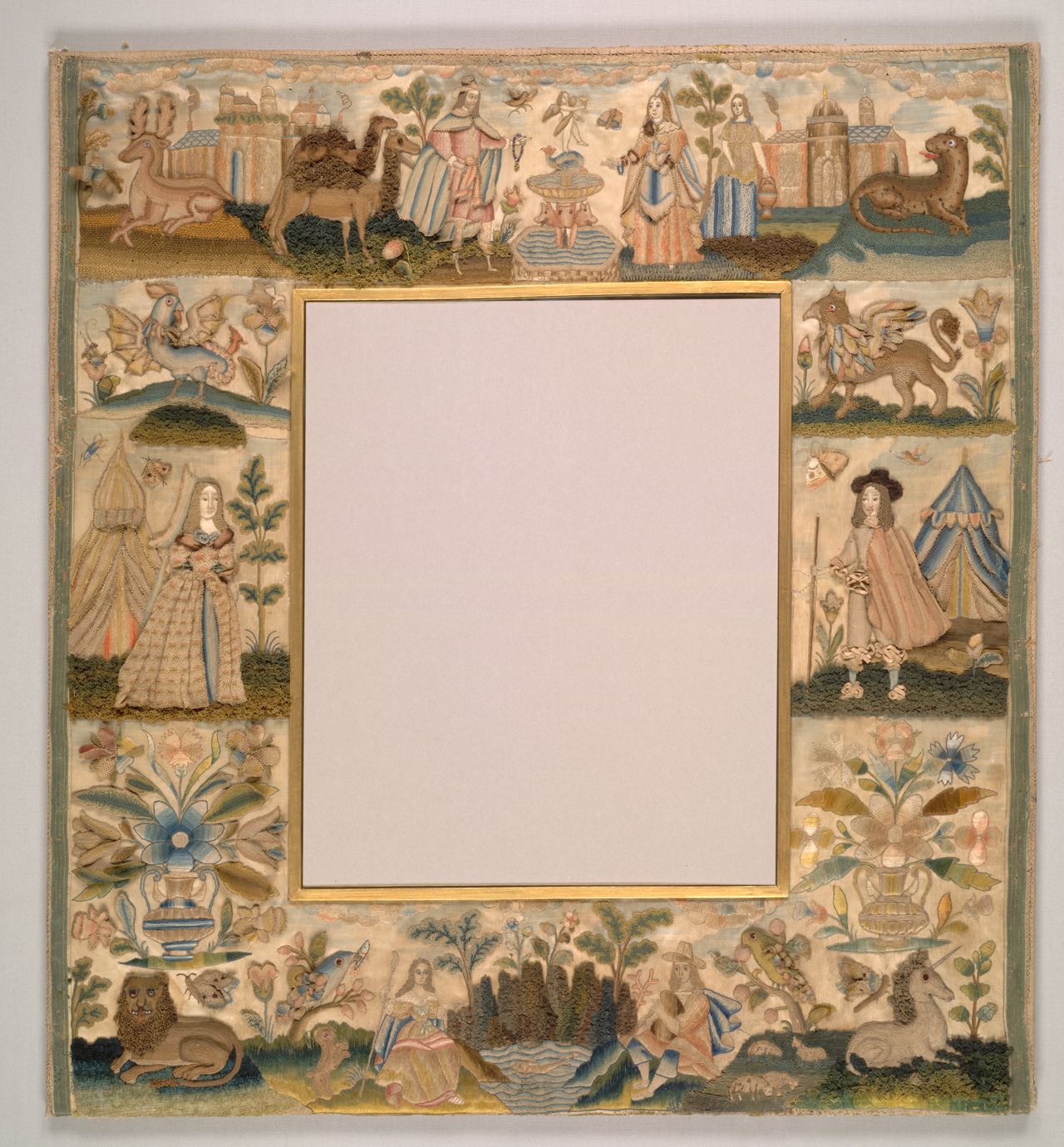A square mirror frame whose border of cream satin is embroidered with small scenes: a woman and a man in seventeenth-century dress, each beside a tent, castles, a camel, a lion, a unicorn, a leopard, a winged dragon and vases of flowers, around an empty central mirror opening.