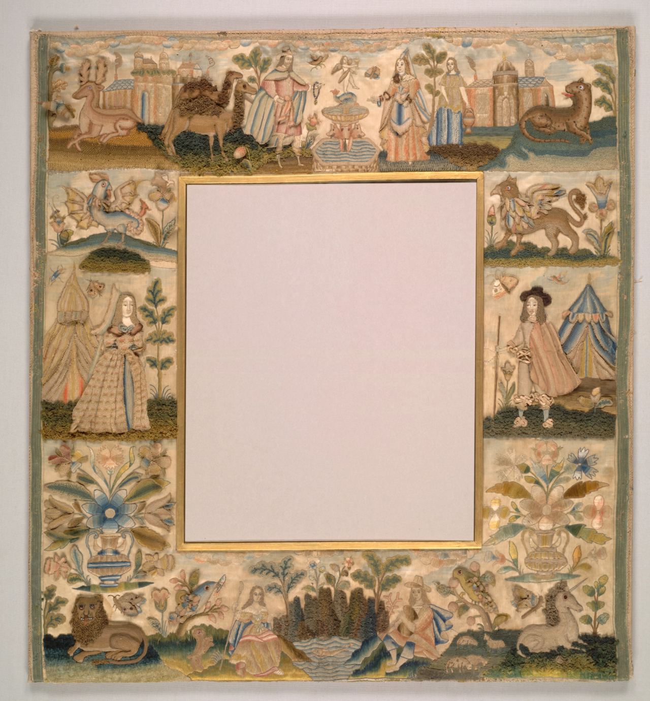 A square mirror frame whose border of cream satin is embroidered with small scenes: a woman and a man in seventeenth-century dress, each beside a tent, castles, a camel, a lion, a unicorn, a leopard, a winged dragon and vases of flowers, around an empty central mirror opening.