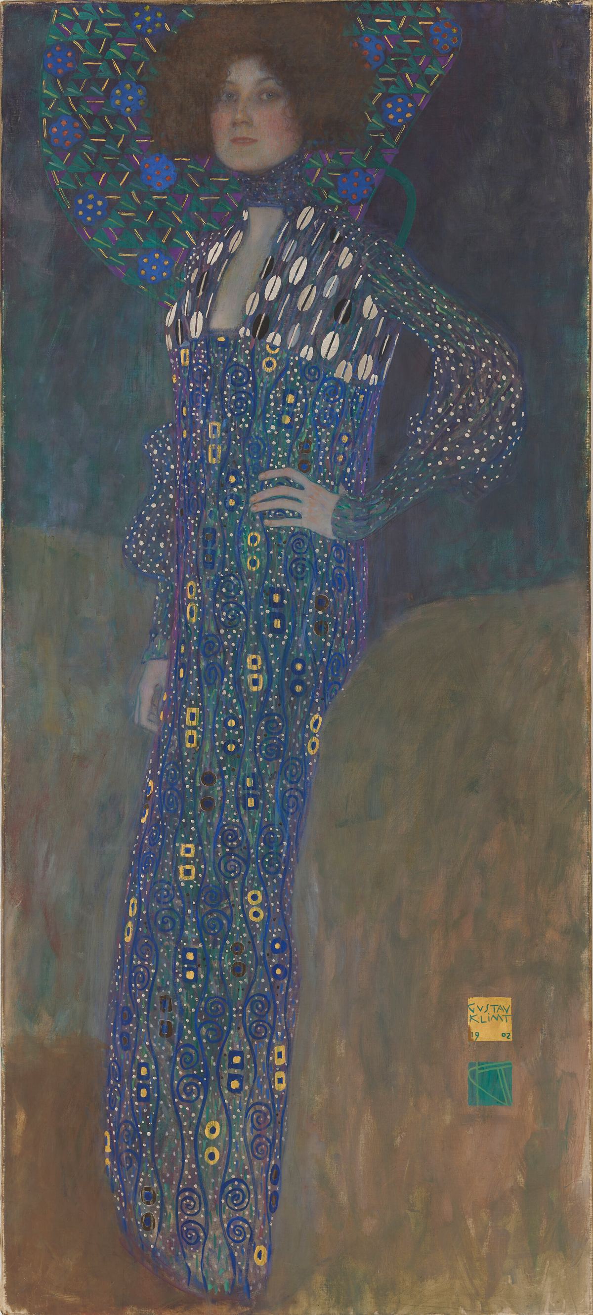 Full-length portrait of a woman with full brown hair standing against a dark blue-green and olive ground, one hand on her hip. She wears a long, narrow, waistless gown covered in blue spirals and small gold and black squares, with a sheer, white-dotted upper bodice and sleeves and a high collar; a large blue-and-green patterned panel rises behind her head like a halo.